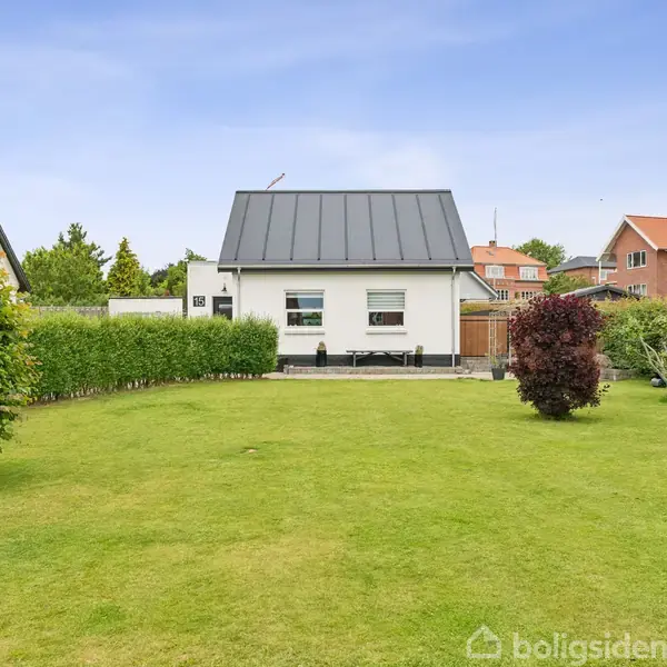 Et hvidt hus med sort tag står i midten af en velplejet have. Buske og træer omgiver huset, der ligger i et beboelseskvarter med lignende bygninger i baggrunden.