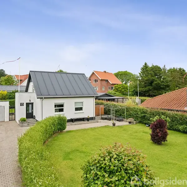Et hvidt hus med sort tag står på en velholdt græsplæne med buske. En indkørsel fører op til huset, der ligger i et grønt boligområde med flere huse i baggrunden.
