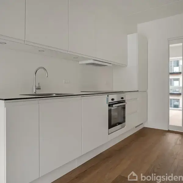 Et moderne køkken med hvide skabe og en indbygget ovn langs væggen. Der er en vask med vandhane. Det åbner sig til et lyst rum med trægulv og vinduer.
