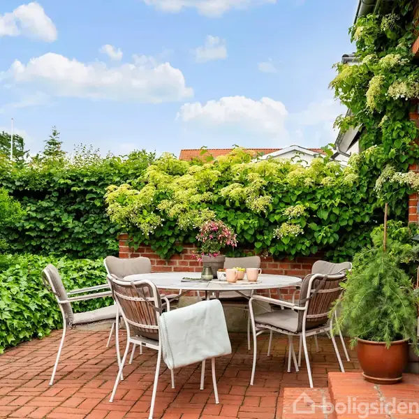 Rundt havebord med stole står på en flisebelagt terrasse ved en murstensbygning. Borden er pyntet med planter og morgenmadselementer, omkranset af grønne buske og klar himmel.