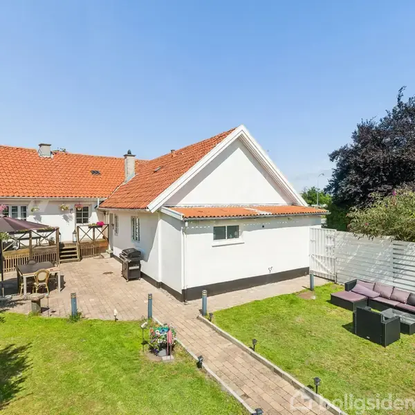 Hus med hvid facade og rødt tegltag står sammen med en solrig terrasse og græsplæne. I haven ses havemøbler og en grill, omkranset af hegn og træer.