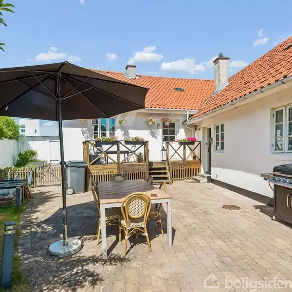 Terrassemøbler stående under en stor sort parasol ved siden af en grill. Omgivet af en murstensbelagt gårdhave og et hvidt hus med rødt tegltag.