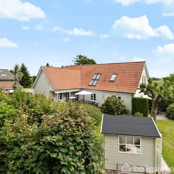 Et hvidt hus med rødt tegltag står i en frodig have. En terrasse med parasol ses foran huset, omringet af buske og træer.