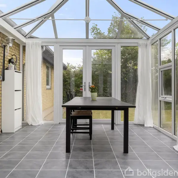 Vinterhave med glasvægge og -loft. Et rektangulært bord med to stole står centralt. Omgivet af flisegulv, gardiner og græsterrasse udenfor.