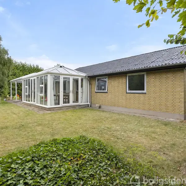 En murstensbygning med sort tag har en tilstødende glasveranda. Den står i en græsplæne omkranset af træer og buske. En legeplads med røde elementer ses til venstre.