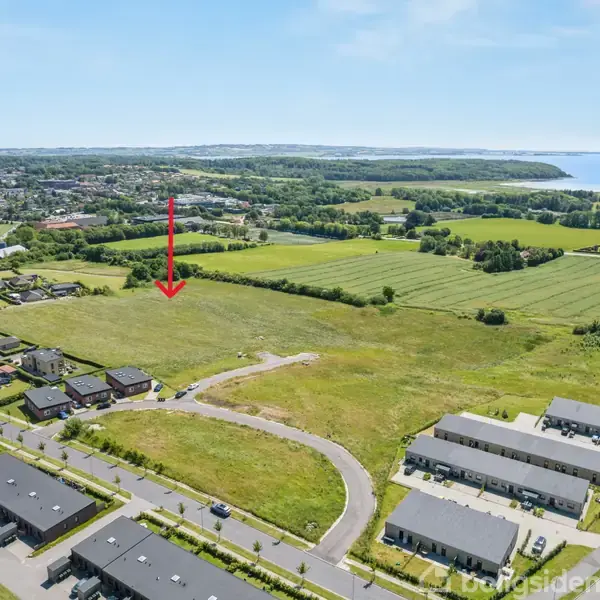 Et stort, åbent markområde markeret med en rød pil, omgivet af beboelseshuse og veje, med en baggrund af grønne marker og en fjord i horisonten.