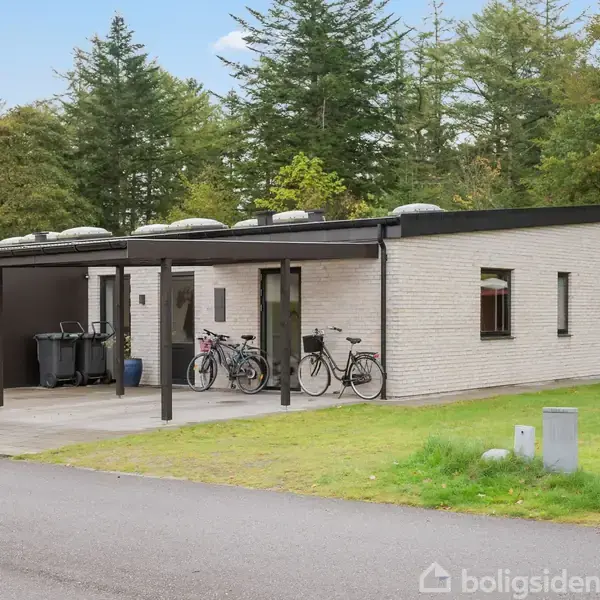 En modern villa med fladt tag står stille i et grønt boligområde. Tre cykler er parkeret foran huset under en sort carport. Grus- og asfaltvej i forgrunden.