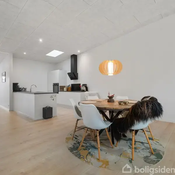 Et rundt spisebord med stole står i et lyst rum med trælaminatgulv. En køkkenø med vask ses i baggrunden, og en orange lampeskærm hænger over bordet.