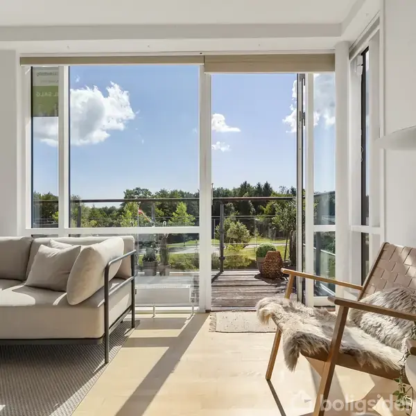 En stue med en beige sofa og lænestol, åbent mod en balkon gennem glasdøre. Jeg hænger en lampe over et træbord, omkranset af en grøn udsigt udenfor.