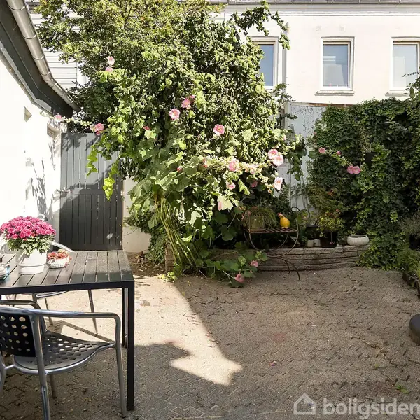 En gårdhave med et sort bord og stol, hvor der står en vase med lyserøde blomster. Området er omgivet af grønne planter og en sammenklappet parasol.