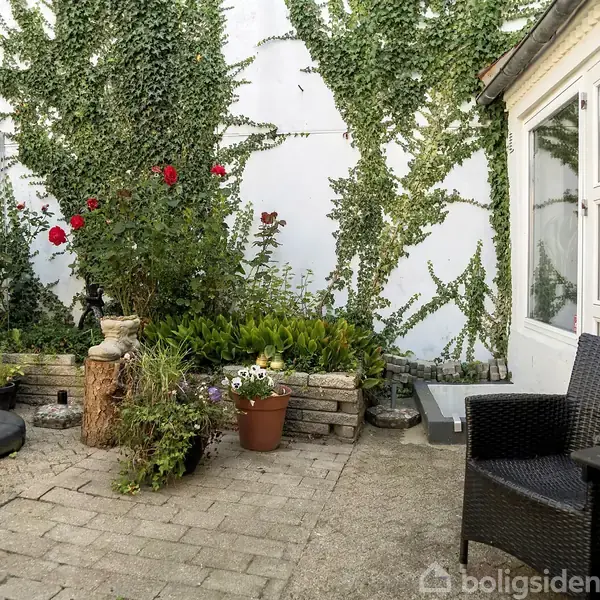 En gårdhave med flisebelægning, hvor en parasol står til venstre. Blomster og grønne planter vokser langs en hvid væg. Til højre er der sorte havemøbler.