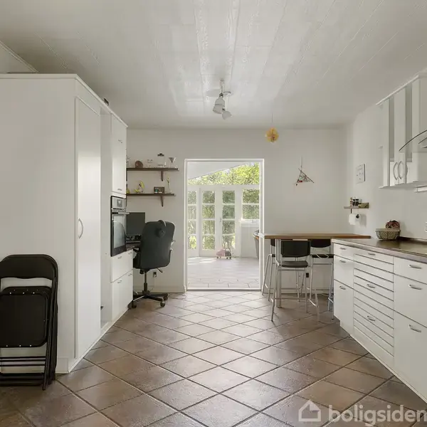 Et funktionelt køkken med hvide skabe og sorte hylder; der er et spisebord ved vinduet. Flisegulv og dør til terrasse med udsigt til en grøn have.