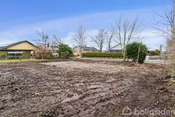 Bar jordgrund, uden beplantning, ligger i et beboelsesområde omgivet af enkelte træer og buske. I baggrunden ses huse og vej under en klar blå himmel.