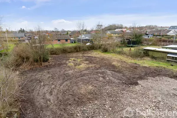 En bar jordgrund ligger stille i et beboelsesområde. Grunden er omgivet af træer og buske, med huse og garager i baggrunden under en delvis overskyet himmel.