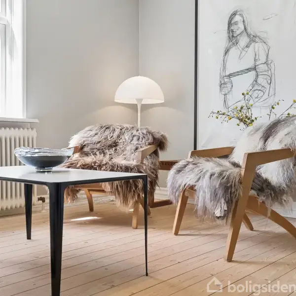 To stole dækket med pelstæpper står ved et lavt bord, hvorpå der er en skål. En gulvlampe lyser ved siden af en stor indrammet skitse. Trægulv og store vinduer i omgivelserne.