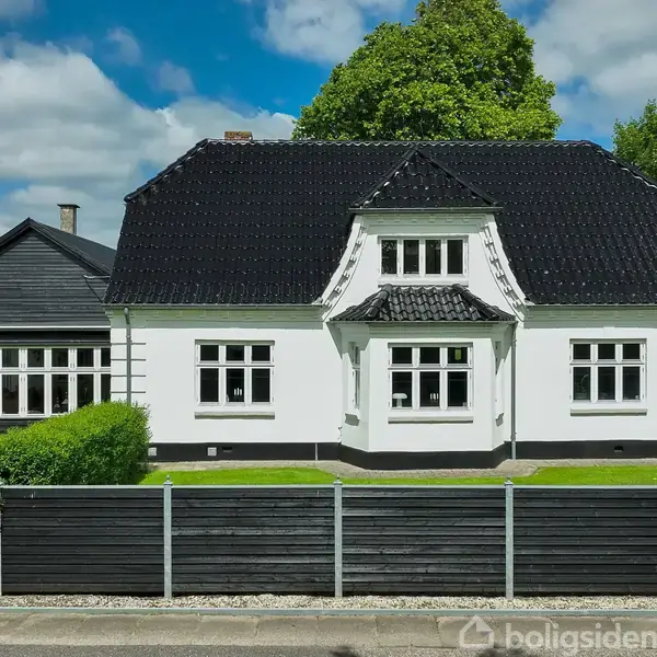 Et hvidt hus med sort tag og sprossevinduer står omgivet af en velholdt have og træer. Et sort stakit adskiller ejendommen fra fortovet foran.