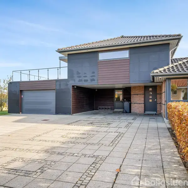 En moderne to-etagers hus med fladt tag, garage og stor indkørsel. Huset er omgivet af græsplæne og hække, og ligger i et villakvarter med træer i baggrunden.