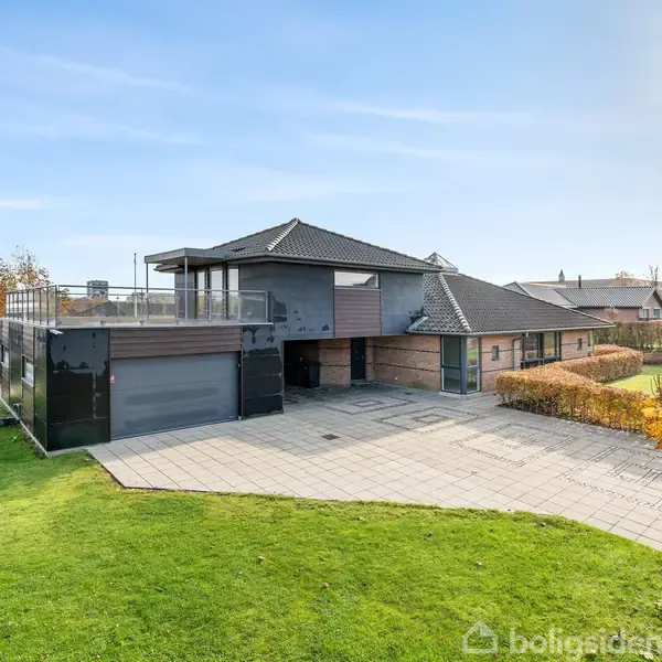 Hus med moderne design står stille på en stor græsplæne i forstæderne. En stor indkørsel fører op til garagen, med et solrigt, klart himmel som baggrund.