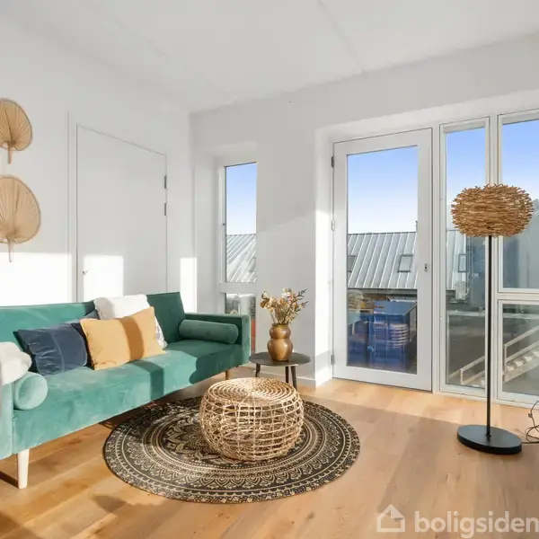 Grøn sofa med farverige puder står i en lys stue. Rund rattanlampe og bord matcher tæppe. Store vinduer giver udsigt til en balkon med klart himmelbaggrund. Ingen tekst.
