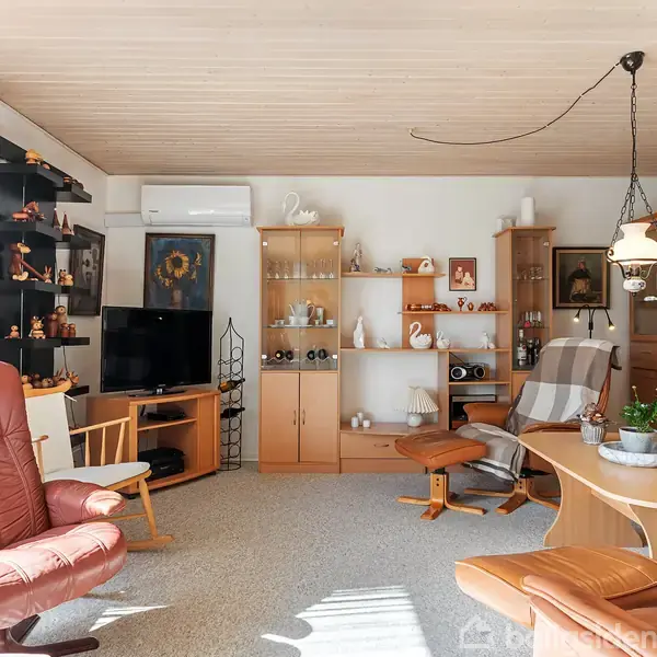 Stue med lænestole og sofabord; reol fyldt med porcelæn, planter og små dekorationer. Et fjernsyn står ved siden af en lænestol. Sollys kommer ind gennem store vinduer.