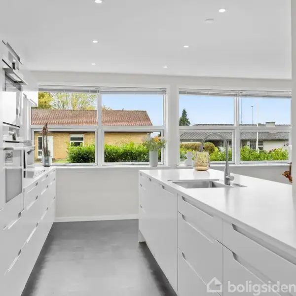 Et moderne køkken med hvide skabe og en stor ø i midten. Et vindue bagved giver udsigt til en forstadsområde med huse og grønne buske.