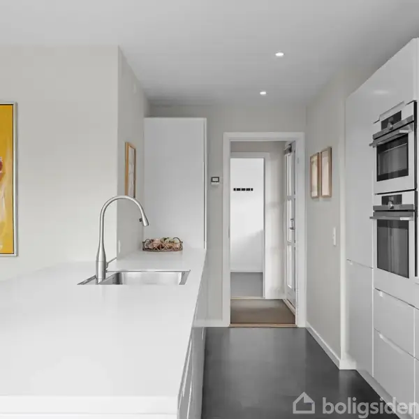 Et moderne køkken viser en hvid bordplade med en vask, flankeret af et farverigt maleri. I baggrunden ses en døråbning og indbyggede ovne ved siden af skuffer.