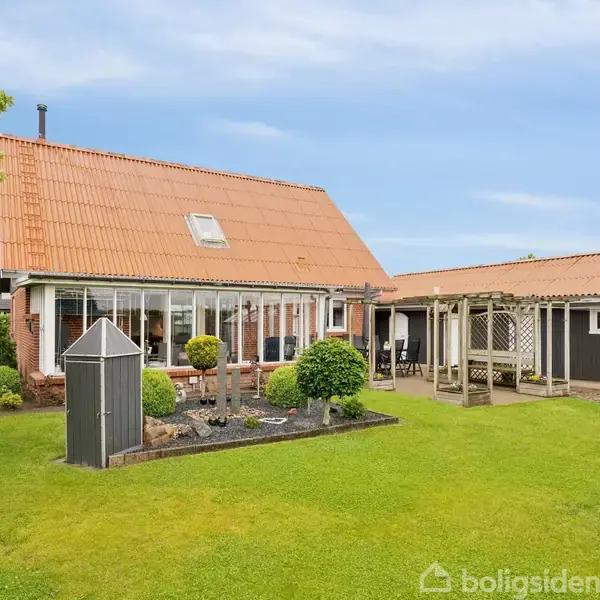 Hus med rødt tegltag og stor glaskarnap står i frodig have med grøn græsplæne og træer. Til højre ses en træterrasse med overdækning.