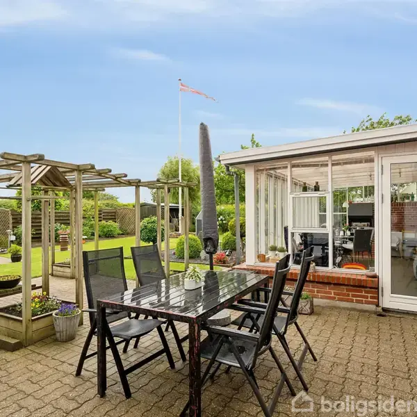 Sort udendørs bord med seks stole står på en flisebelagt terrasse. Solseng og grønne planter omgiver området, mens en flagstang ses i baggrunden ved siden af et glashus.