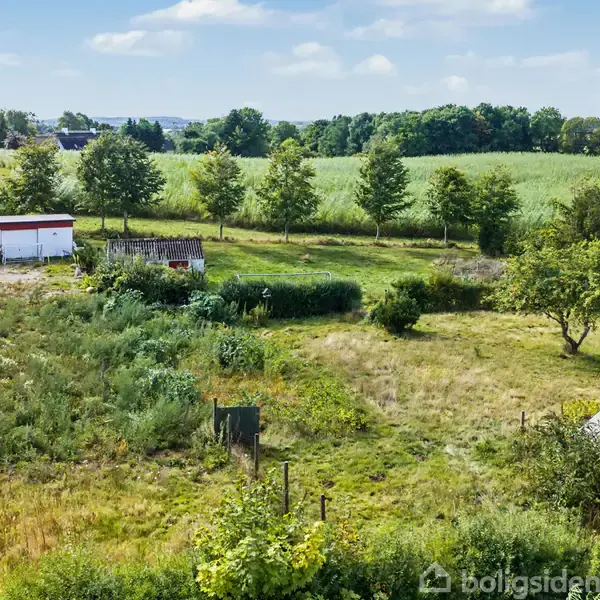 Grunden er et åbent, grønt areal med ukontrolleret vegetation. Små skure står til venstre. Vejen løber langs kanten, og markerne udgør baggrunden. Skyfri, solrig himmel.