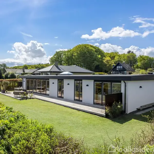 En moderne, hvid villa står stille omgivet af en velplejet have med grønne buske. Sollys belyser husets terrasse. Bagved ses en skovbevokset bakke og en klar blå himmel.