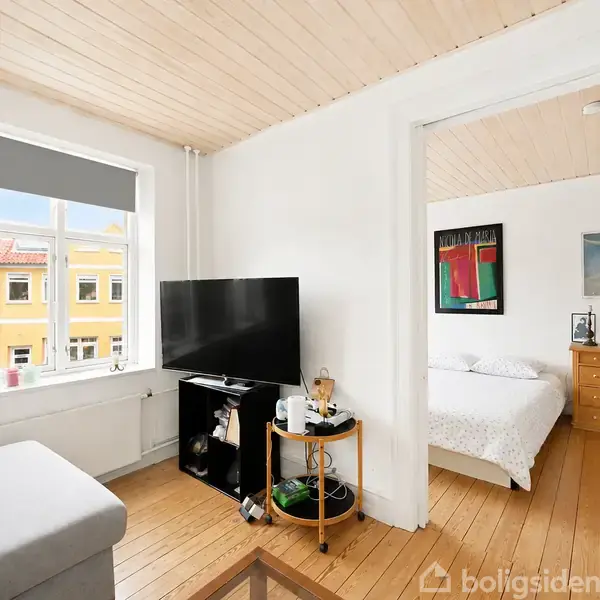 En stue med grå sofa har et TV stående på et bord ved vinduet med solskin. I baggrunden ses et soveværelse med dobbeltseng og kommode.