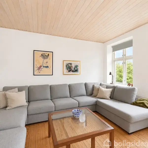 Grå sofa står i hjørnet af et lyst rum med trægulv, flankeret af en træbord med vaser. Væggen har to billeder, og en lampe hænger fra loftet.