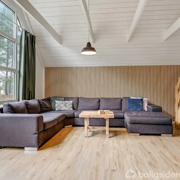 En stor, grå hjørnesofa står i et lyst rum under en skråloft, flankeret af store vinduer og træpaneler. En trappe fører opad. En træbord står foran sofaen.