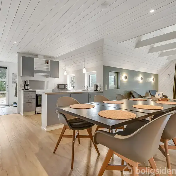 Et moderne spisestue- og køkkenområde med et langt bord dækket med dækkeservietter og stole, i et rum med hvidmalede vægge og lofter. Store vinduer tillader naturligt lys.