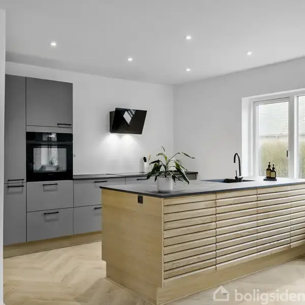 Et moderne køkken med en central træ-ø med planter og en mørk bordplade. I baggrunden ses grå køkkenelementer med indbyggede apparater. Store vinduer slipper lys ind.