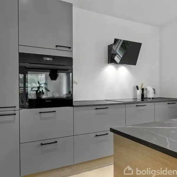 Gråt køkken med moderne indbyggede skabe og skuffer, ovn og emhætte. På bordpladen står en kaffemaskine og en brødrister. Murstensgulv i lys træfarve omkranser rummet.