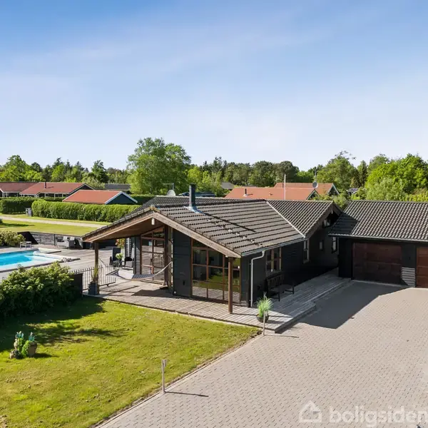 En moderne enfamiliebolig med fladt tag står ved siden af en indkørsel og swimmingpool omgivet af en velplejet have og flere omkringliggende huse under en klar himmel.