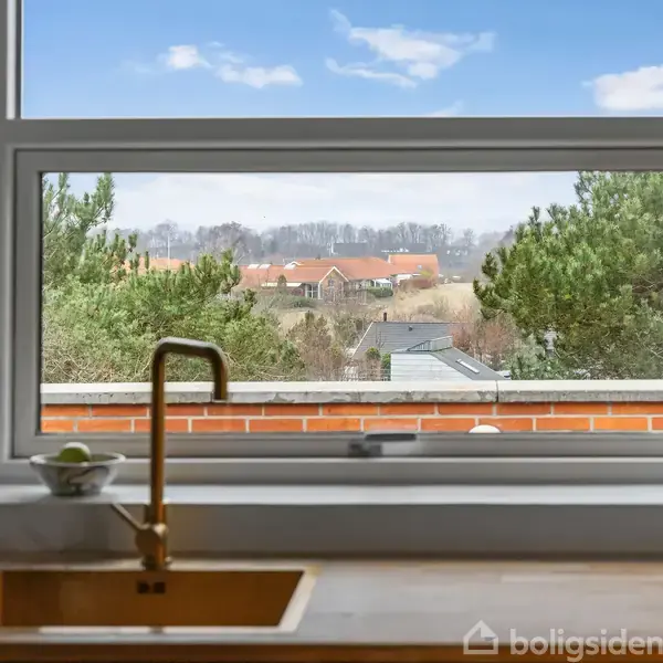 En guldfarvet vandhane over en køkkenvask med udsigt til landskab gennem et stort vindue. Træer og bygninger ses i det fjerne under en klar blå himmel.