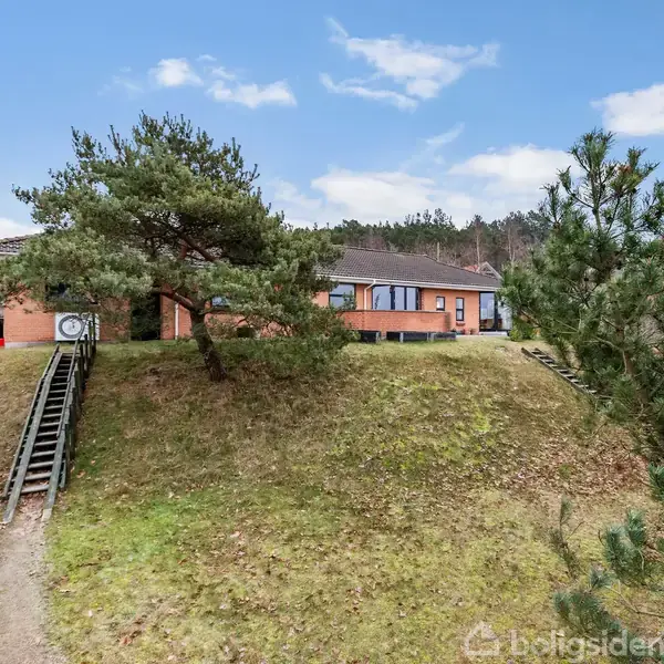 Et rødt murstenshus står på en græsklædt bakke med en stor trappe foran. Omgivet af træer og en klar blå himmel i baggrunden.
