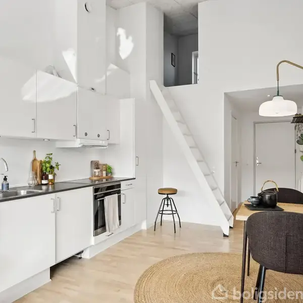 Et lyst, moderne køkken med hvidt design, hvor en stige fører op til en hems. Til højre står et spisebord med to stole, omgivet af dekorative elementer.