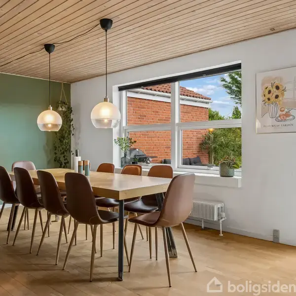 Spisebord omgivet af otte stole, står i en lys spisestue med grøn væg. To runde lamper hænger over bordet, og et vindue viser en udsigt til en murstensbygning. En plakat med "fil de fer vases" hænger på væggen.