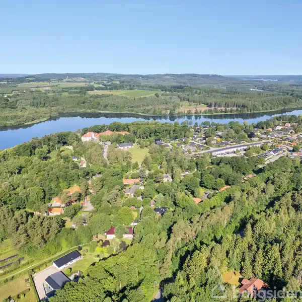 Landskab med flod bugter sig gennem tæt skovklædt område. I forgrunden ses huse og en lille by med veje og bebyggelse, omgivet af frodig natur under blå himmel.