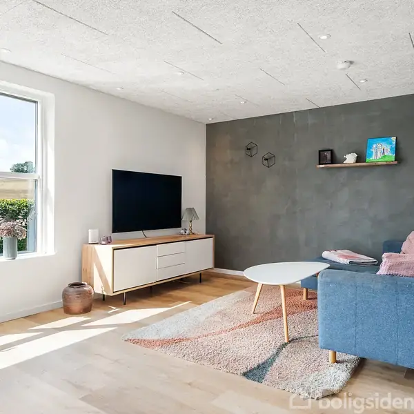 En stue med en blå sofa, et rundt sofabord og et TV på en skænk. Væggen er grå med hylder, og der er et stort vindue med udsigt til en hæk.
