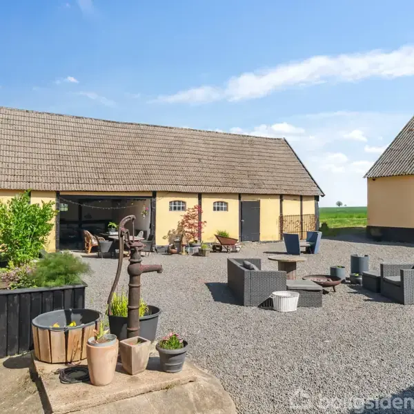 Gårdsplads med grusbelægning, hvor der står en gammel vandpumpe og udemøbler. Omgivet af gule landbrugsbygninger med træ- og plantedejligheder, under en klar blå himmel i landligt område.