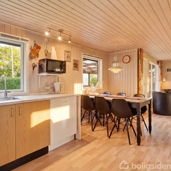 Køkken i sommerhusstil med træloft og -gulv, overskab og vask. Lang spisebord med sort stole i midten. Store vinduer giver naturligt lys. Vægure og udendørs udsigt.