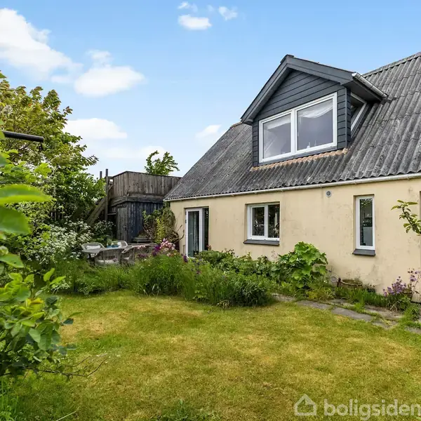 Hus med skråtag og kvistvindue, står i en frodig have med græs og buske. Terrassen til venstre giver et hyggeligt udemiljø. Skyfri himmel i baggrunden.