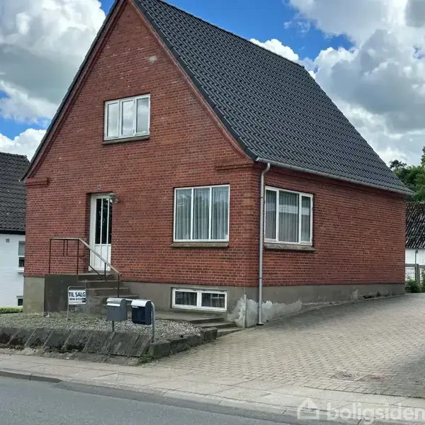 Et rødt murstenshus med sorte tagsten står til salg. Huset har hvide vinduer og en dør med en lille trappe foran. Omgivet af en flisebelagt sti og et par postkasser. Skilt: "Til salg."