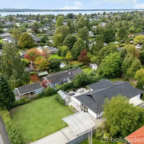 Huset, der er hovedfokus, står stille omgivet af en velplejet have med en carport. Det ligger i et grønt boligområde med flere huse, træer og udsigt til vand i baggrunden.