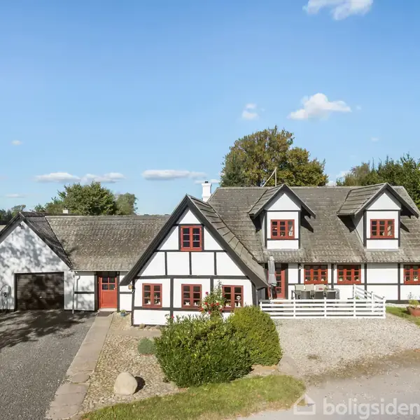 Et hvidt bindingsværkshus med røde vinduer står i en solrig landlig have. Grusvej og buske omgiver bygningen, mens en klar blå himmel ses i baggrunden.
