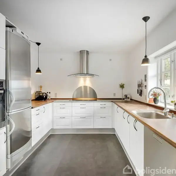 Et moderne køkken viser en vinkelopstilling med hvide skabe, stålkomfur og opvaskemaskine. Lyset strømmer ind gennem vinduer med udsigt til grønne planter udenfor.